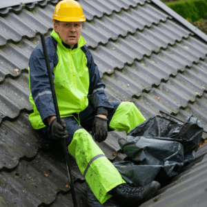 a worker in high-visibility rain gear and a hard hat clears debris from a commercial roof’s drainage system, emphasizing the importance of maintaining gutters and downspouts to prevent water buildup and leaks.