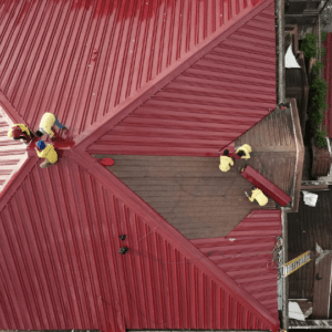 an aerial view of workers inspecting and repairing a large red metal roof, illustrating the importance of post-holiday roof inspections to identify and address any winter-related damage.