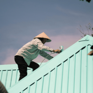 a worker applies a protective coating to a light blue metal roof under clear skies, demonstrating the importance of pre-summer roof maintenance to protect against UV damage and heat exposure.