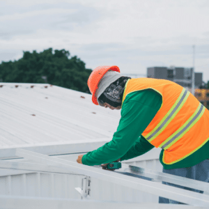 a roofing contractor in a high-visibility vest and hard hat inspects and secures a metal commercial roof, emphasizing the necessity of regular maintenance to prevent structural damage and costly repairs.