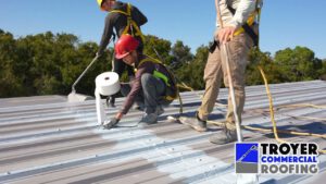 Commercial roofing crew in safety harnesses applying reinforcing fabric during a metal roof restoration