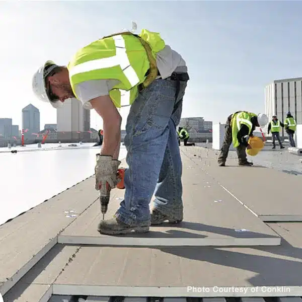 A commercial roofer wearing safety gear uses a power drill to secure insulation boards on a flat rooftop.