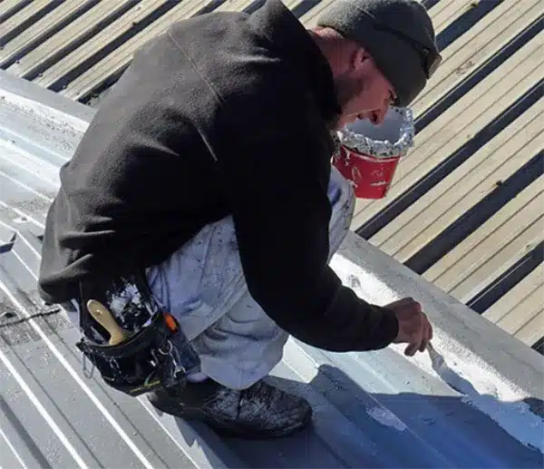 A contractor kneels on a corrugated metal roof, brushing coating along a seam near a supply bucket.