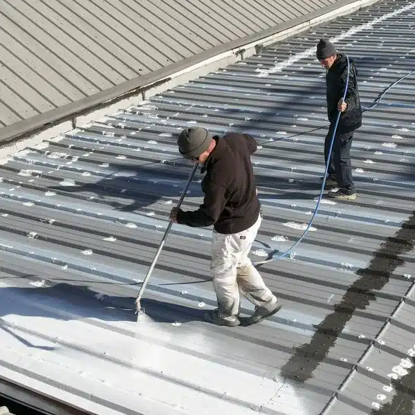 Two roofers apply a white protective coating to a corrugated metal commercial roof using a spray wand