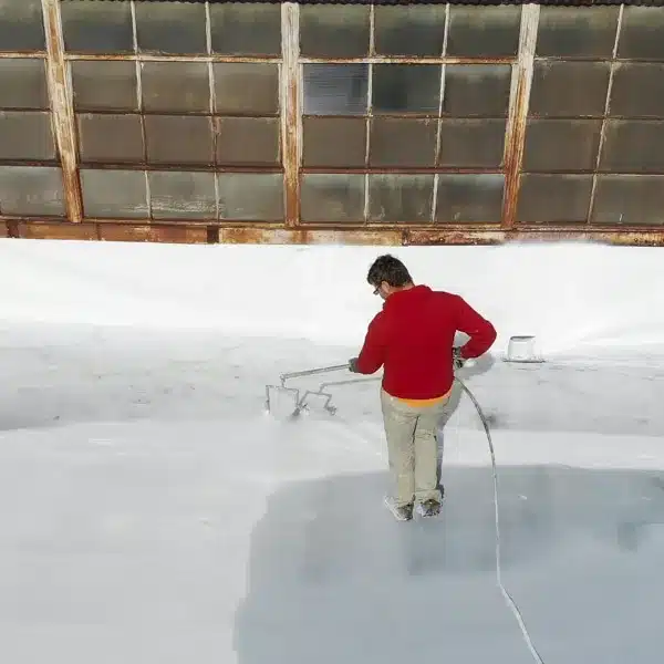 A worker wearing a red jacket sprays a white protective coating onto a flat commercial roof.